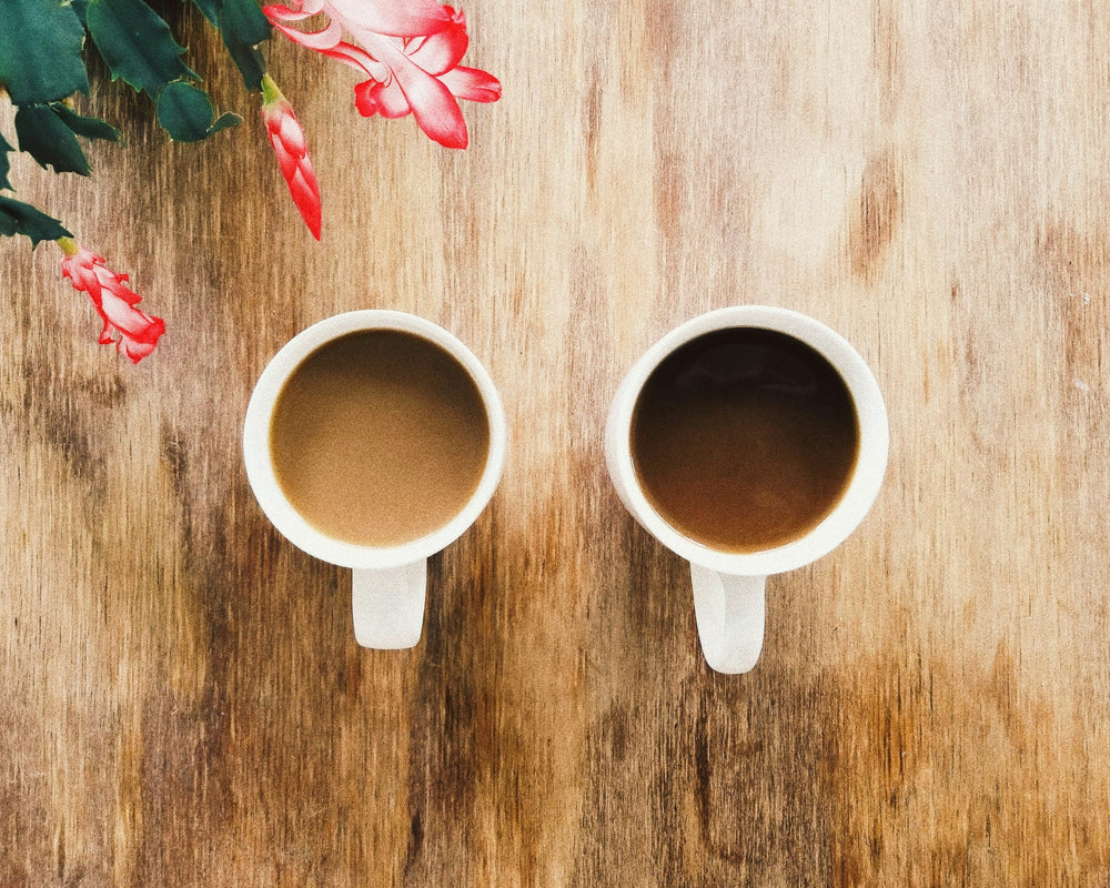 Two ceramic cups of coffee on a wooden table beside flowers.